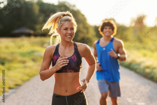 Cute sporty couple running in nature on the sunny summer day. Selective focus on blonde woman with ponytail.