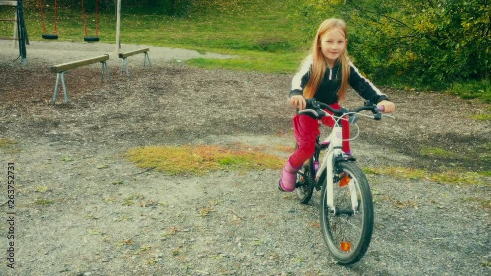 A small child wearing a red dress rides her bicycle towards the camera and smiles as she goes out of frame. A small child wearing a red dress rides her bicycle towards the camera and smiles as she goes out of frame.