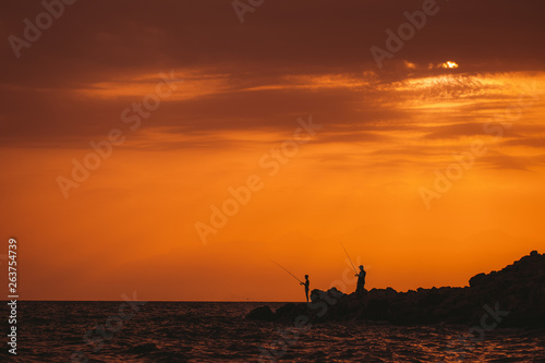 Two black silhouettes of fishermen with rods standing on seashore isolated at orange sunset sky background. Horizontal color photography.