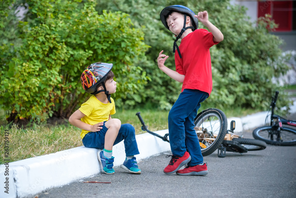 Fototapeta premium little boy fallen off of his bike. Unhappy boy sitting on asphalt looking his knee. His friend helping him to stand up and ride again