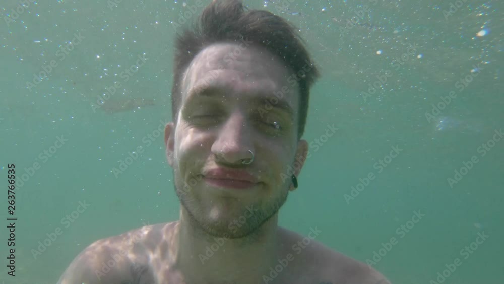 Man in crystal clear ocean water with people on a white sandy beach lined by lush green tropical landscape behind him. Guy holding camera goes underwater to relax and cool off from the Thailand heat.