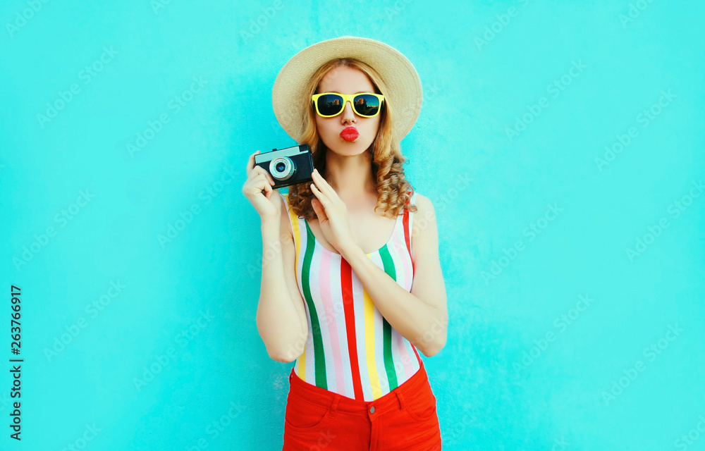 Colorful young woman holding retro camera, blowing red lips sends air kiss in summer straw hat having fun on blue wall background Colorful young woman holding retro camera, blowing red lips sends air kiss in summer straw hat having fun on blue wall background