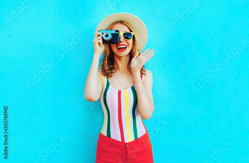 Colorful happy smiling young woman holding retro camera in summer straw hat having fun on blue wall background