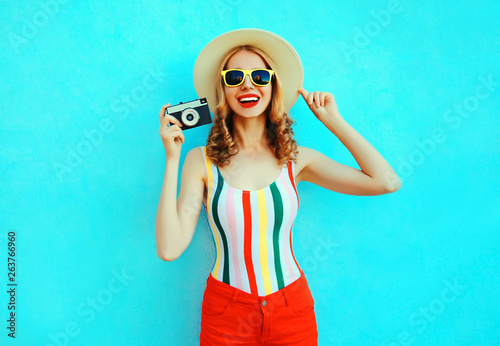 Colorful happy smiling young woman holding retro camera in summer straw hat having fun on blue wall background