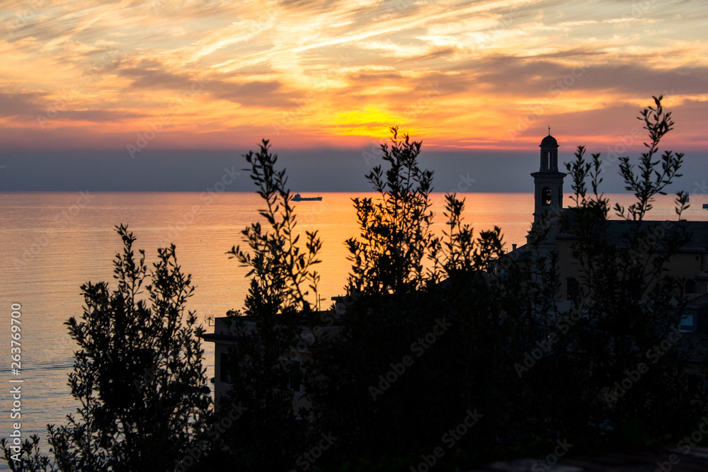 Beautiful yellow and red sunset on cloudy sky near the Mediterranean sea marine of Genoa Boccadasse beach. View from the hill, the sea and a ship with tree on a foreground and tower on the right side.