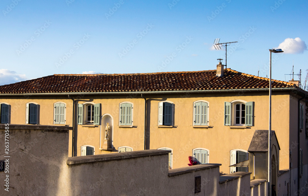 Magnificent French facade of a residential building with shutter windows and tile brown roof in sun light, fence on the foreground and blue cloudless sky above. Summer mood.