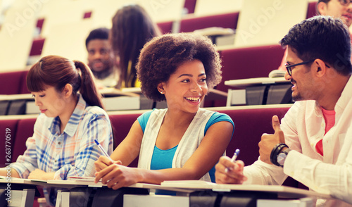 education, high school, university, learning and people concept - group of international students with notebooks writing in lecture hall