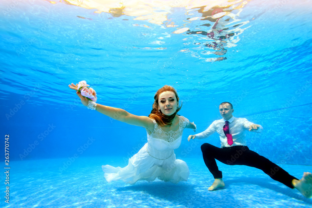 Bride in white dress and groom swim and pose underwater on camera at the bottom of the pool in clear blue water. Portrait. Horizontal orientation