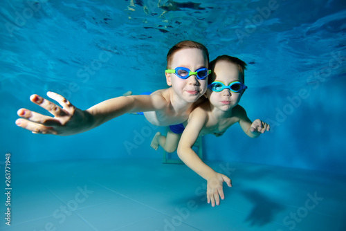 Happy kids, two little boys, swimming underwater in the pool on a blue background. They hug and pose for the camera. Portrait. Close-up. Underwater photography. Horizontal view