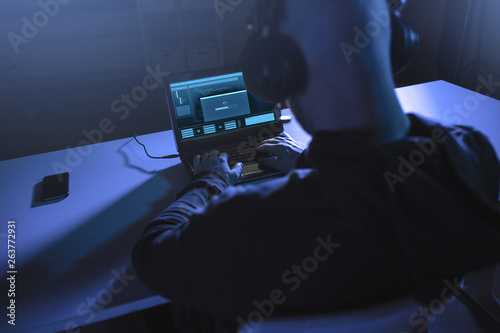cybercrime, hacking and technology concept - male hacker in headphones with progress loading bar on laptop computer screen using virus program for cyber attack in dark room