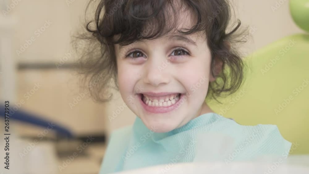 Cute little girl is sitting on a dentist's chair and smiling, showing her teeth to camera. Girl has an appointment with a dentist.