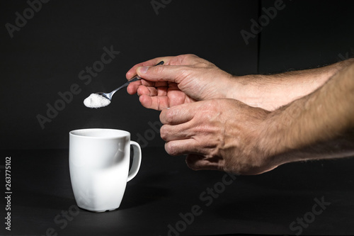 Male hand pouring white sugar on a white mug, isolated over black.