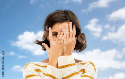 people concept - young woman in striped pullover closing face with hands and looking by one eye through her fingers over blue sky and clouds background