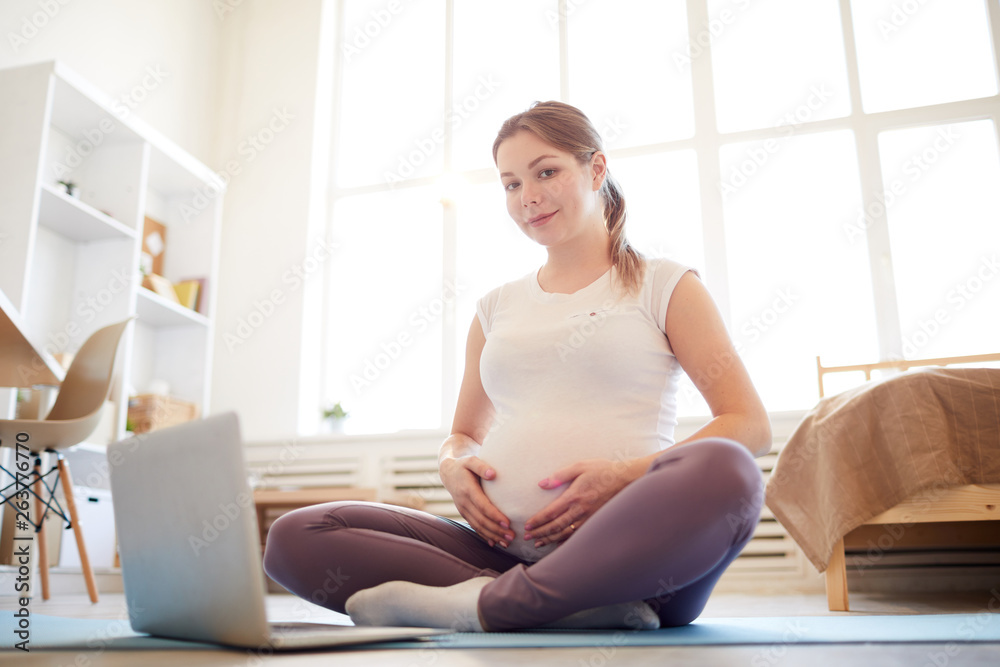Full length portrait of young pregnant woman sitting on yoga mat with laptop and smiling at camera, copy space Full length portrait of young pregnant woman sitting on yoga mat with laptop and smiling at camera, copy space