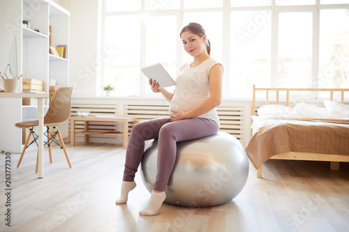 Slika na platnu Full length portrait of pregnant woman sitting on fitness ball and using digital