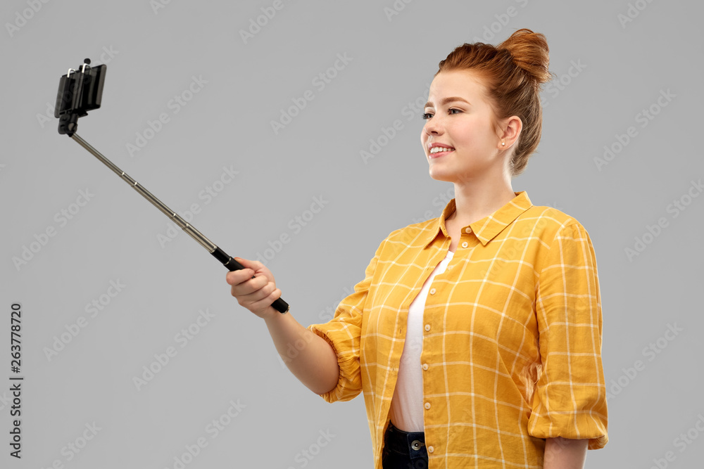 technology and people concept - smiling red haired teenage girl in checkered shirt taking picture by smartphone on selfie stick over grey background technology and people concept - smiling red haired teenage girl in checkered shirt taking picture by smartphone on selfie stick over grey background