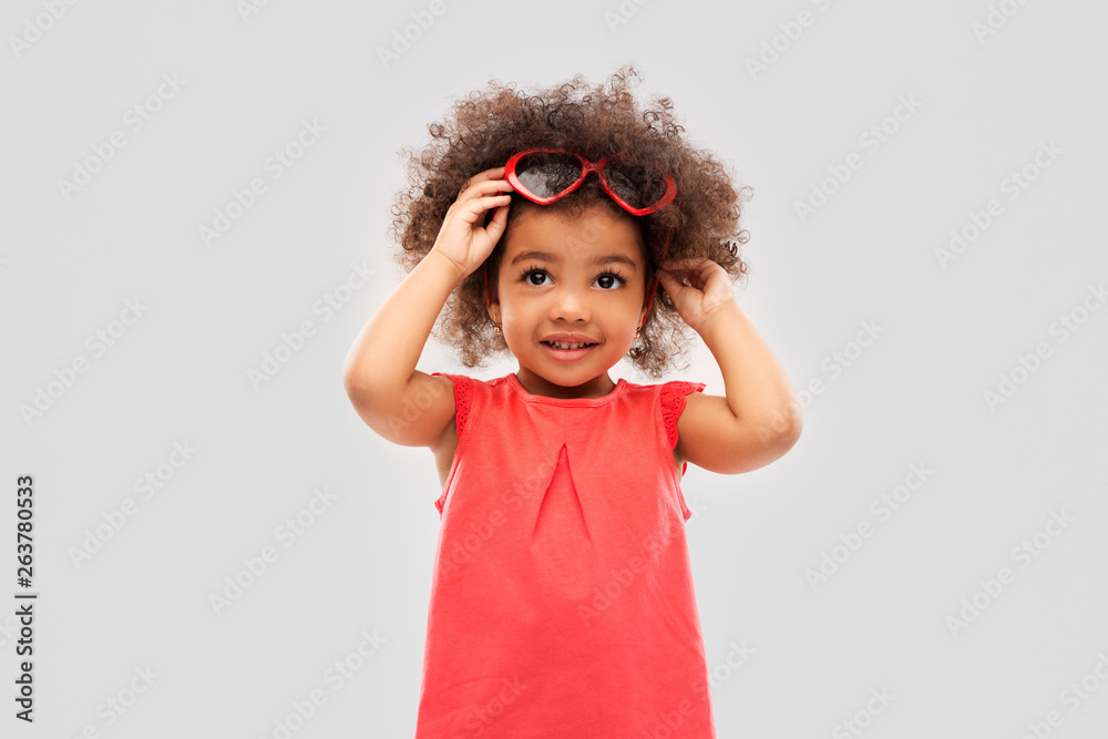 childhood, valentine's day and summer concept - happy little african american girl with heart shaped sunglasses over grey background childhood, valentine's day and summer concept - happy little african american girl with heart shaped sunglasses over grey background