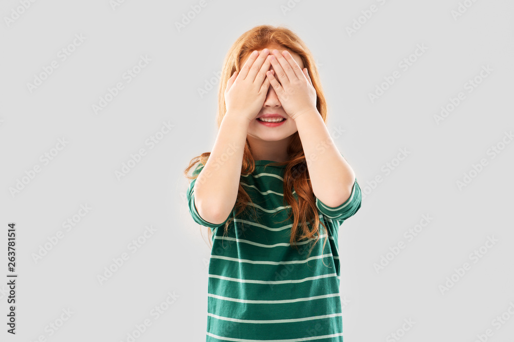 childhood, seeing and vision concept - smiling red haired girl in green striped shirt with eyes closed by hands over grey background childhood, seeing and vision concept - smiling red haired girl in green striped shirt with eyes closed by hands over grey background