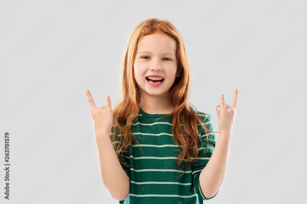 childhood, gesture and people concept - smiling red haired girl in green striped shirt showing rock hand sign over grey background childhood, gesture and people concept - smiling red haired girl in green striped shirt showing rock hand sign over grey background