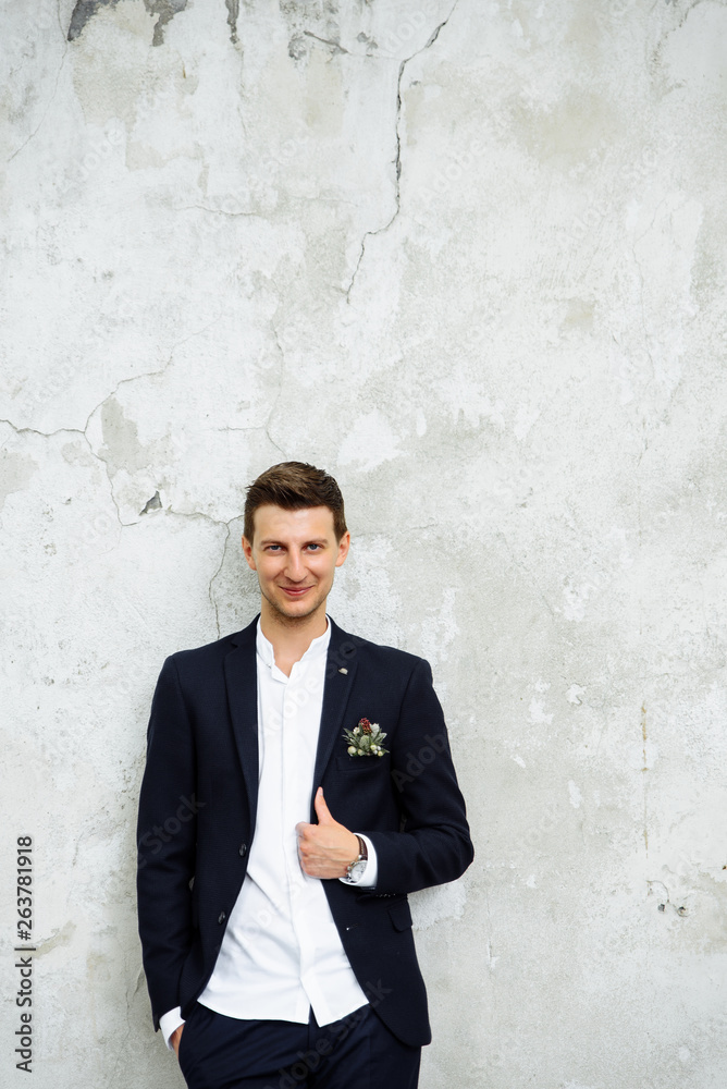 Portrait of a handsome young groom in a stylish modern suit, standing against a white wall and smiling Portrait of a handsome young groom in a stylish modern suit, standing against a white wall and smiling