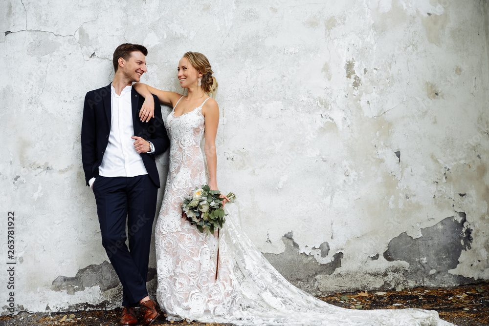 Luxury newlywed couple on the background of a white wall. Bride in a classic long wedding dress, the groom in expensive styled suit