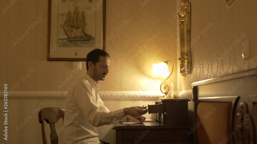 Man in white shirt sitting in a dark room with a table light takes out of a wooden desk drawer his pen and is getting ready to write. Man in white shirt sitting in a dark room with a table light takes out of a wooden desk drawer his pen and is getting ready to write.