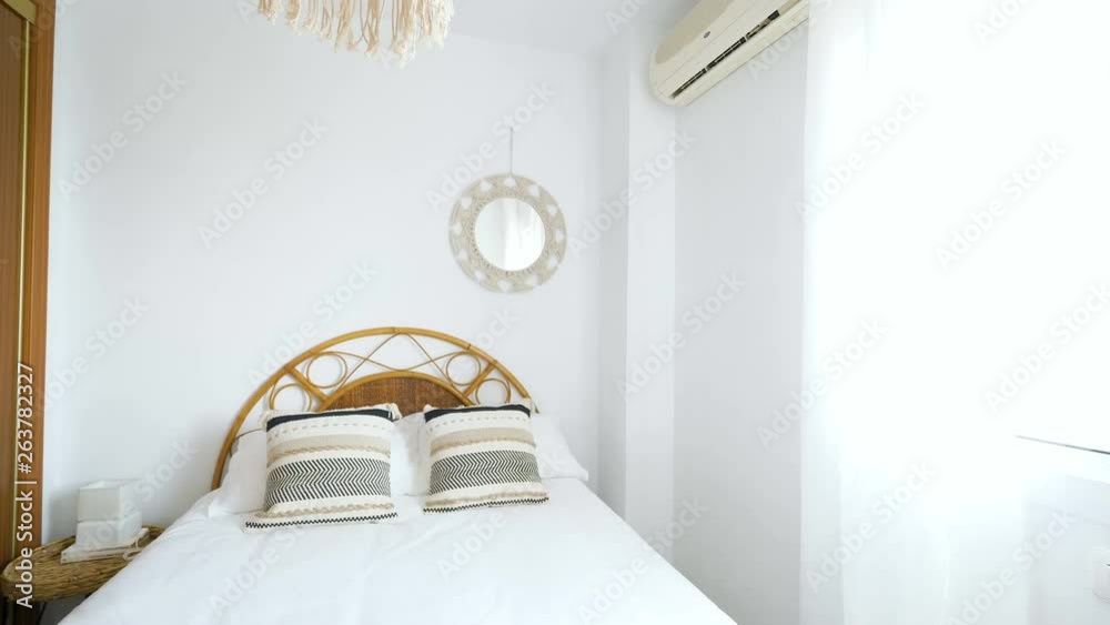 Comfortable hotel bedroom with rattan headboard with natural fabric cushion, macrame mirror, fiber nightstand and a big window with view.Holiday destination apartment with boho contemporary style.