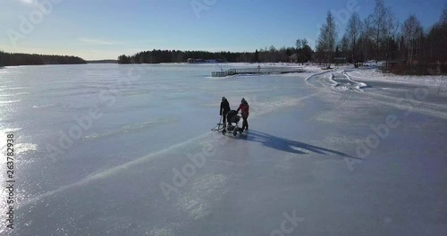 Aerial, young woman with baby stroller and older woman with kick sled walking on frozen lake on a beautiful sunny winter day photo