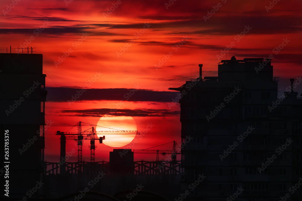 Citysacpe silhouette of buildings and construction site cranes with big bloody fiery red sun at dusk on background. Scenic industrial landscape