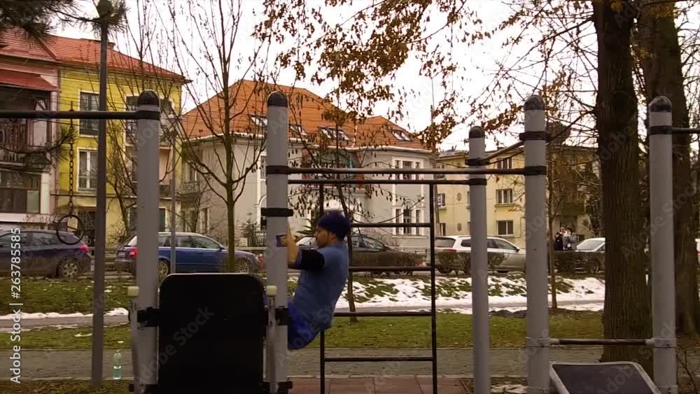 Man in blue swinging between bars of different heights on a piece of exercise equipment near the street in Cluj Central Park, Romania. Homes and cars in the background as a man exercises. Slow motion. Man in blue swinging between bars of different heights on a piece of exercise equipment near the street in Cluj Central Park, Romania. Homes and cars in the background as a man exercises. Slow motion.