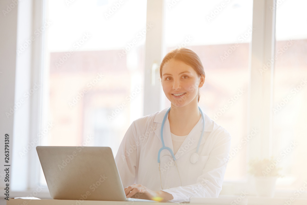Portrait of young female doctor smiling at camera while working in office lit by warm sunlight, copy space Portrait of young female doctor smiling at camera while working in office lit by warm sunlight, copy space