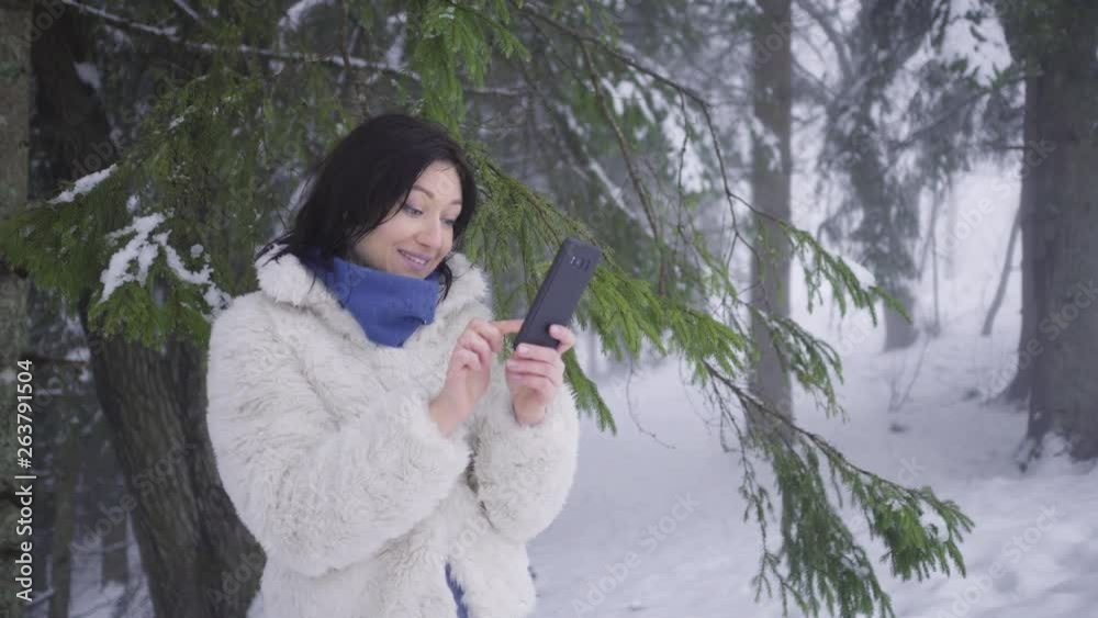 Attractive woman in white fur coat and blue scarf making selfie with black smartphone by spruce branches - snowy mist in winter forest Attractive woman in white fur coat and blue scarf making selfie with black smartphone by spruce branches - snowy mist in winter forest