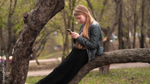 Young girl sitting on tree branch and looking at smartphone side shot photo