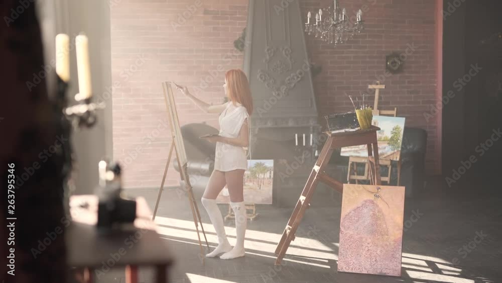 Wonderful young red-haired girl is standing in her workroom in only white transparent shirt and painting. The room is made in a loft style. Wonderful young red-haired girl is standing in her workroom in only white transparent shirt and painting. The room is made in a loft style.