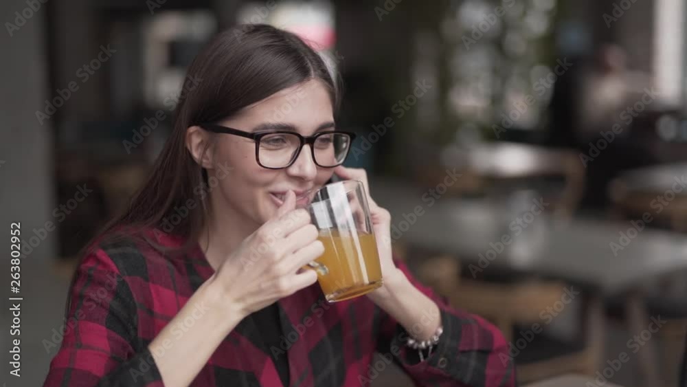 Attractive young dark-haired woman in a glasses is sitting in a cafe indoors, talking on the phone, drinking an orange tea and smiling. Attractive young dark-haired woman in a glasses is sitting in a cafe indoors, talking on the phone, drinking an orange tea and smiling.