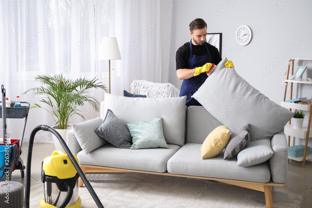 Male janitor cleaning sofa in room - obrazy, fototapety, plakaty 
