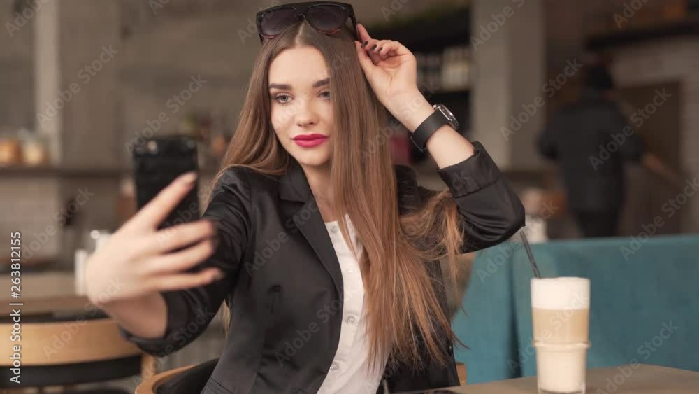 Beautiful Girl With Long Brown Hair and Red Lipstick Sitting in Cafe for Coffee Break and Takes Selfies With Smartphone While She Holds Sunglasses On Head With Other Hand. Blurred Background Beautiful Girl With Long Brown Hair and Red Lipstick Sitting in Cafe for Coffee Break and Takes Selfies With Smartphone While She Holds Sunglasses On Head With Other Hand. Blurred Background