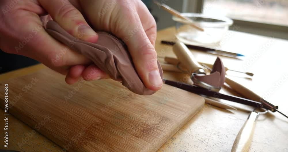 Slide across the hands of an artist sculpting with brown modeling clay on a wooden table with tools strewn about the art studio.
