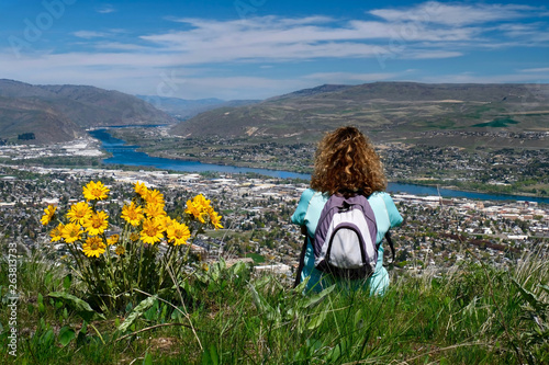 Woman with backpack sitting on the top of mountain enjoying scenic views of city and river. Spring wild flowers on hills. Wenatchee. Washington. United States photo