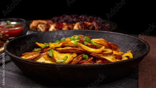 Fried potato chips with greens in a pot, in front of homemade sausages and stewed cabbage, on a black rustic background