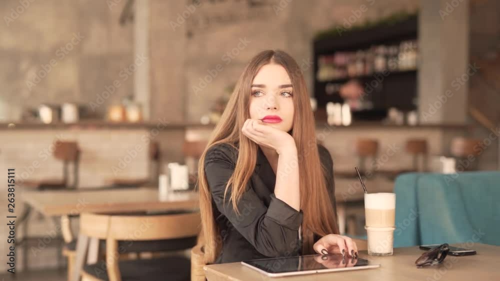 Young Woman With Red Lipstick and Brown Hair Sitting na Cafe Holding Her Head In Hand Looking Outside Deeply In Thoughts. Coffee Tablet and Sunglasses on Table Young Woman With Red Lipstick and Brown Hair Sitting na Cafe Holding Her Head In Hand Looking Outside Deeply In Thoughts. Coffee Tablet and Sunglasses on Table