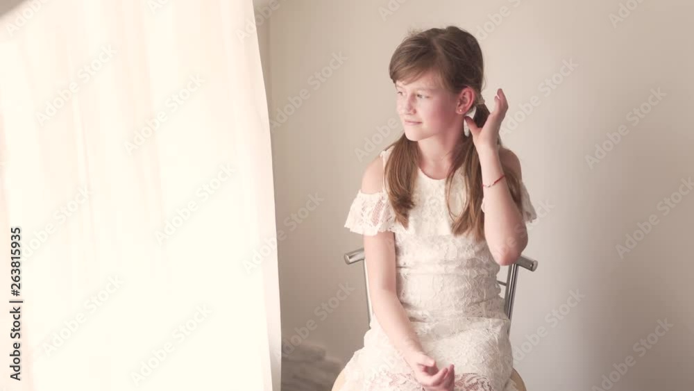 Child Portrait of Girl Sitting in Chair By Window on Daylight in White Dress Moving Her Pigtals to Look Perfetcly and Looks Through Window. Camera Close Up.