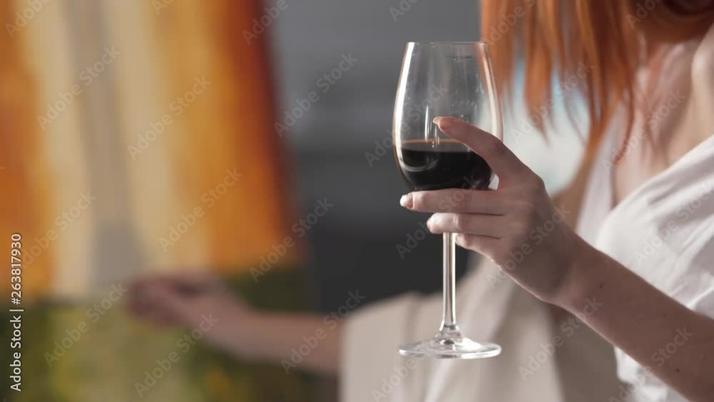 A glass of red wine in focus in the foreground is held by a woman who paints an out of focus canvas in the background with her other hand.