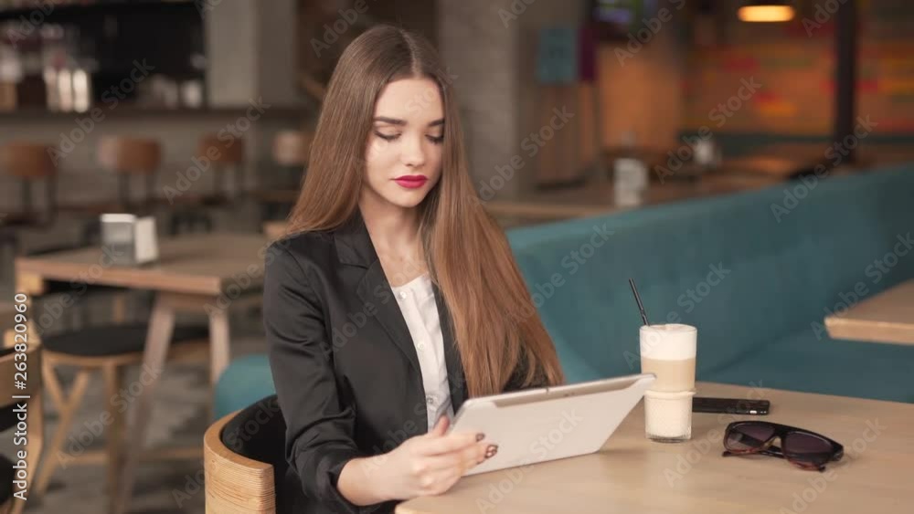A gorgeous lady with long brunette hair, pink lips and lovely face wears long black jacket, white blouse and beige trousers sits in the restaurant. She is busy and focused entirely on her tablet. A gorgeous lady with long brunette hair, pink lips and lovely face wears long black jacket, white blouse and beige trousers sits in the restaurant. She is busy and focused entirely on her tablet.