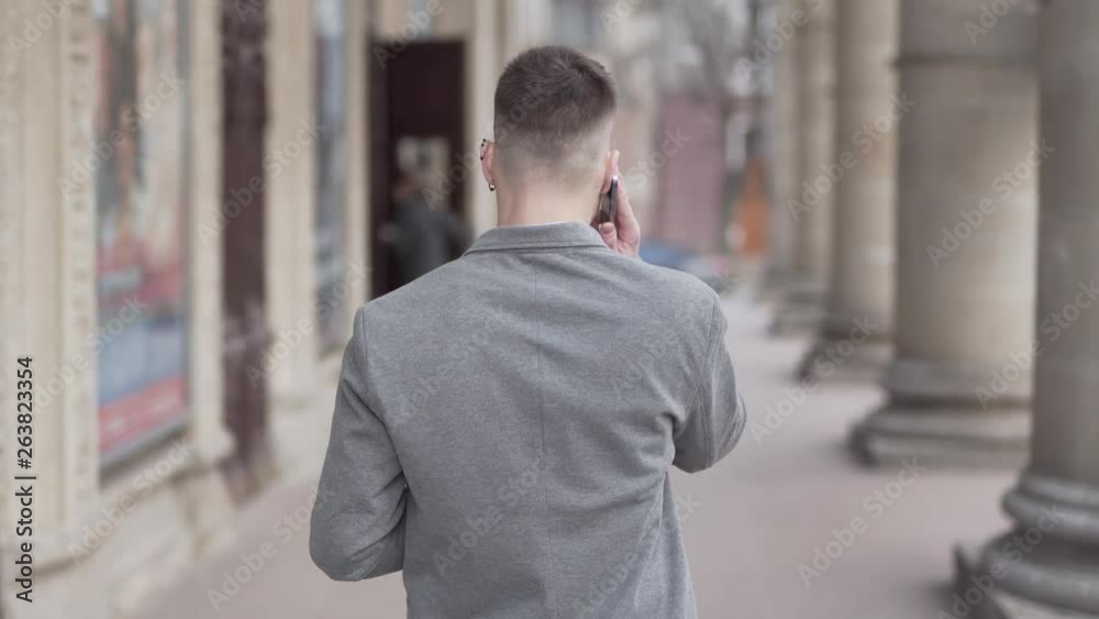 Young Male In Grey Jacket Walking By Old Building and Talking On Smartphone. Camera on His Back in Follow Mode. Blurred Background. Young Male In Grey Jacket Walking By Old Building and Talking On Smartphone. Camera on His Back in Follow Mode. Blurred Background.