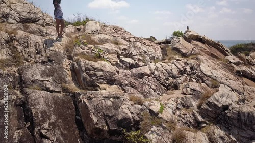 Aerial: A drone rises its alttitude and reveals a girl standing at steep rocks and stones looking around the tropical landscsape, at some point the camera slowly pans to the left. Thailand. photo