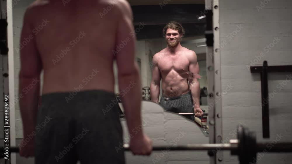Topless young bearded male showing off and training biceps hard by doing bicep curls with a barbell in front of a broken mirror in an old, rusty Arnold era gym (Gold's Gym) close-up Topless young bearded male showing off and training biceps hard by doing bicep curls with a barbell in front of a broken mirror in an old, rusty Arnold era gym (Gold's Gym) close-up