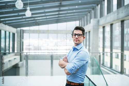 A portrait of young businessman standing indoors in an office, arms crossed.