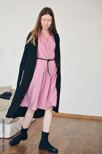 Beautiful girl without makeup in pink dress with black belt under black linen coat in black shoes on home interior background