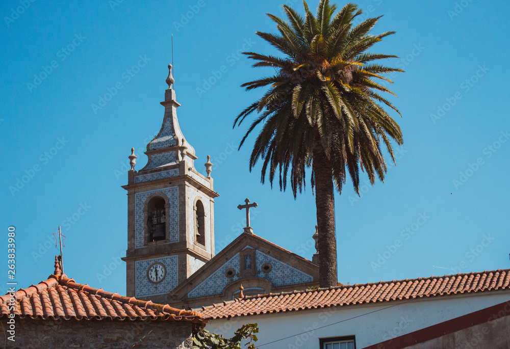 Traditional portuguese church with azulejos tiles with palm tree and old buildings. Mediterranean religious landmark. Catholic tower with cross and bell. Sunny day in beautiful village, Europe. 